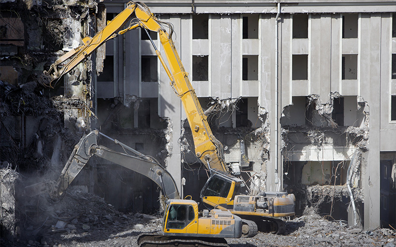 Two large yellow excavators demolish a concrete building, creating rubble.